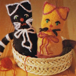 May include: Two handmade knitted cat dolls in a woven basket. One cat is black with white accents and striped arms, the other is yellow with pink accents. Both have embroidered faces and are tied with yarn bows. A red ball of yarn sits nearby.