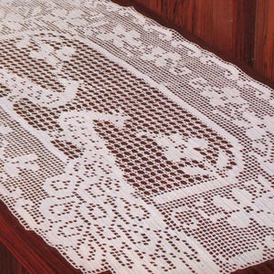 May include: A white, rectangular, crocheted lace table runner with a floral and geometric pattern. The runner is placed on a wooden surface, showcasing its intricate design and delicate texture. The edges are scalloped.