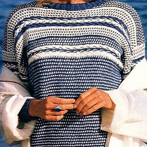 May include: A blue and white knitted jumper with a round neck. The jumper has a textured pattern with horizontal stripes and a cable knit design. The person is wearing a white jacket and a blue ring.