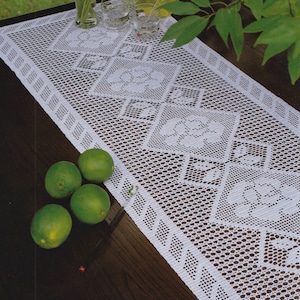 May include: White crocheted table runner with a floral pattern. The rectangular runner features a repeating design of square floral motifs and diamond borders. Four green limes are on the table.