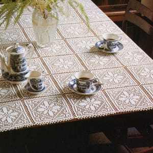 May include: A detailed shot of a dining table set for tea. The table is covered with a white, open-weave lace tablecloth. Tea cups and saucers, a teapot, and a creamer are arranged on the table. The tea set has a blue and white design.