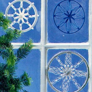 May include: Four decorative circular ornaments, each with a unique design, hang in front of a blue background. Two are white, one is blue, and one is silver. The ornaments are made of a woven material, resembling snowflakes or starbursts. A green Christmas tree branch is in the foreground.
