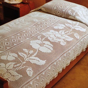 Könnte beinhalten: Ein cremefarbenes, gehäkeltes Bettwäsche-Set mit Bettdecke und Kissenbezug. Die Bettdecke hat ein Blumendesign mit detaillierten Mustern und einem gewellten Rand. Der Kissenbezug spiegelt das Design der Bettdecke wider.