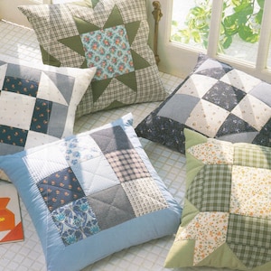 May include: A collection of handmade patchwork throw pillows in various designs and colors. The pillows feature geometric patterns, floral prints, and plaid fabrics in shades of blue, green, and neutral tones. The pillows are arranged on a light-colored surface.