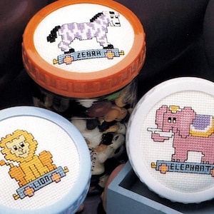 May include: Cross-stitch jar lids featuring a zebra, lion, and elephant. The zebra is purple and white, the lion is yellow, and the elephant is pink. Each animal is on a blue and white base with its name.