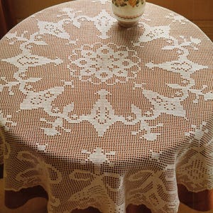 May include: A round, cream-colored crocheted tablecloth with an intricate floral and geometric design. The tablecloth is draped over a round table with a brown border. A small, decorative vase sits on top.
