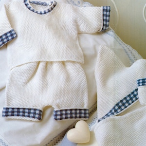Puede incluir: Un conjunto de ropa de bebé de punto color crema, que incluye una parte superior y pantalones cortos, con ribetes a cuadros azules y blancos en las mangas, el cuello y los puños de los pantalones cortos. También hay una toalla con capucha a juego. Un jabón en forma de corazón está en primer plano.