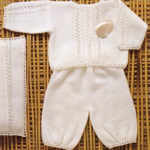 May include: A white knitted baby outfit, including a long-sleeved top and matching pants. The outfit features decorative openwork details along the sleeves, neckline, and center of the top. A small, light-colored seashell rests on the top. The outfit is displayed on a light brown, woven surface.