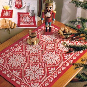 May include: A red and white embroidered Christmas table runner with a geometric snowflake pattern. The image also shows matching placemats, napkins, and a nutcracker figurine, creating a festive holiday setting.