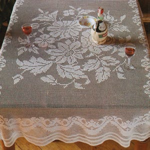 May include: A beige crocheted tablecloth with a floral design covers a wooden table. Two wine glasses with red liquid are on the table, along with a small bottle and a dish. The tablecloth has a scalloped edge.