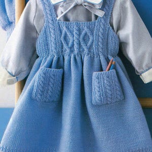 Könnte beinhalten: Ein hellblaues Strickkleid mit Zopfmusterdetails an Oberteil und Taschen. Das Kleid hat einen quadratischen Ausschnitt und zwei Vordertaschen. Ein weißes Hemd mit Kragen und Schleife ist darunter. Stifte sind in einer Tasche sichtbar.