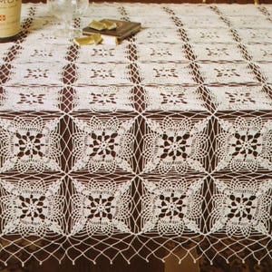 Könnte beinhalten: Ein weißes, gehäkeltes Tischtuch mit einem sich wiederholenden floralen und geometrischen Muster. Das komplizierte Design weist quadratische Motive mit detaillierten floralen Zentren auf, die einen zarten und eleganten Look erzeugen. Das Tischtuch liegt auf einer braunen Oberfläche.