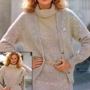 May include: A light gray knit ensemble featuring a turtleneck sweater, cardigan, and skirt. The outfit includes a matching belt with a gold buckle and a small pink flower pin. The image also includes the text "18 NATASCHA/SABA" and "19/19a".