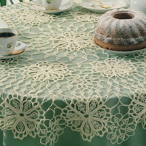 May include: A round table with a light green tablecloth and a cream-coloured, openwork lace overlay. A bundt cake, teacups, saucers, and a creamer are on the table. The lace overlay features a floral design.