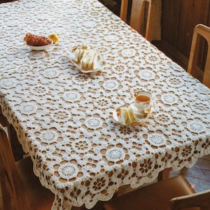 May include: A rectangular, cream-colored, crocheted tablecloth with a floral pattern covers a wooden table. The table is set with small plates of food and a teacup. The tablecloth is approximately 72 inches long and 48 inches wide.