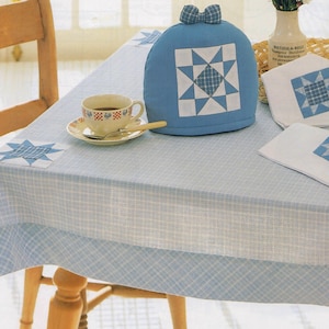 Puede incluir: Un mantel patchwork azul claro y blanco con un calentador de té y servilletas a juego. El calentador de té tiene un lazo en la parte superior y un patrón de estrella. Una taza y un platillo están sobre la mesa. La mesa está puesta con una silla de madera.