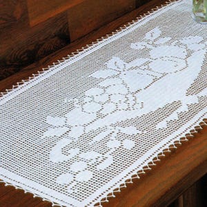 Puede incluir: Camino de mesa rectangular blanco de ganchillo con un diseño de vid. El camino de mesa está colocado sobre una superficie de madera oscura. Los bordes del camino de mesa tienen un ribete decorativo festoneado. Un jarrón de cristal con flores es visible en el fondo.