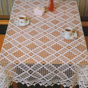 May include: A white, open-weave lace tablecloth with a geometric diamond pattern. The tablecloth is draped over a wooden table, with two teacups and saucers visible. A small pink container and a red vase are also on the table.