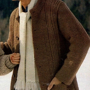 Op de afbeelding: Een bruin, lange mouwen vest met een geribbelde gebreide ontwerp. Het vest heeft een knoopsluiting en zakken. Een crèmekleurige sjaal met franjes wordt gedragen met het vest. De persoon draagt een zonnebril en een blauwe spijkerbroek.