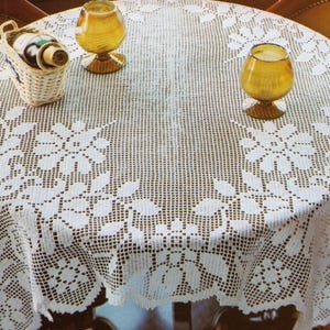 Puede incluir: Un mantel redondo blanco de ganchillo con un diseño floral y de hojas. Dos vasos de color ámbar y una pequeña cesta con botellas están sobre la mesa. El mantel está drapeado sobre una mesa redonda.