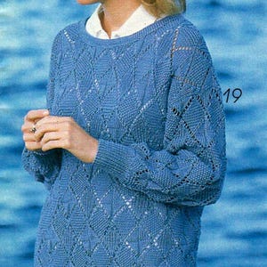 May include: A light blue knitted sweater with a diamond pattern. The sweater has long sleeves and a crew neck. The number "19" is visible on the sleeve. The sweater is worn over a white collared shirt.
