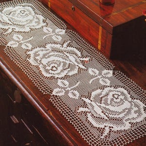 以下が含まれることがあります： 白いオープン織りのレースのテーブルランナー。バラの花柄が繰り返されています。 ランナーは長方形で、手作りのように見え、複雑なディテールが施されています。 バラはランナーの長さに沿って目立つように表示されています。