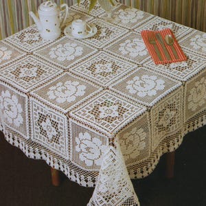 May include: A square, off-white crocheted tablecloth with a floral pattern. The tablecloth is draped over a dark brown table. A matching teapot, teacup, and red and white checkered napkin with silverware are on the table.