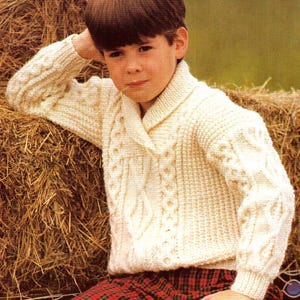 Può includere: Un maglione bianco a treccia con un colletto. Il maglione presenta un tradizionale motivo a treccia e viene indossato da un giovane.