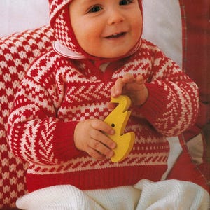 May include: A baby wearing a red and white patterned knit sweater, hat, and pants. The baby is holding a yellow wooden toy. The sweater and hat have a white geometric design. The baby is smiling.