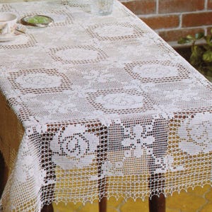 Op de afbeelding: Een wit, gehaakt tafelkleed met een bloemmotief bedekt een houten tafel. Een kop en schotel, en een klein schaaltje staan op het tafelkleed. De randen van het tafelkleed hebben een geschulpte rand, wat een delicate uitstraling geeft.