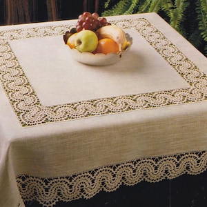 May include: A cream-coloured tablecloth featuring a detailed lace border. A silver bowl filled with fruit, including grapes, an apple, and an orange, sits on the table. The tablecloth has a square design with a detailed lace pattern.