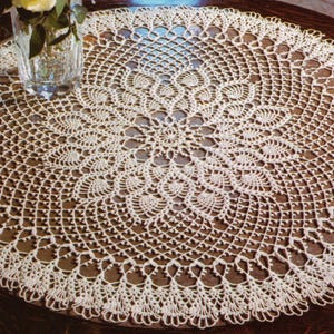 Puede incluir: Tapete de ganchillo color crema con un diseño calado detallado. La pieza circular presenta un motivo floral central rodeado de patrones repetidos y un borde festoneado. Un jarrón de cristal con una flor está en el fondo.