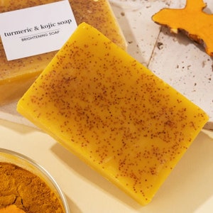 Puede incluir: Una barra de jab&oacute;n de c&uacute;rcuma y kojic, un jab&oacute;n aclarador, de color amarillo con peque&ntilde;as motas marrones. El jab&oacute;n est&aacute; sobre una superficie blanca.