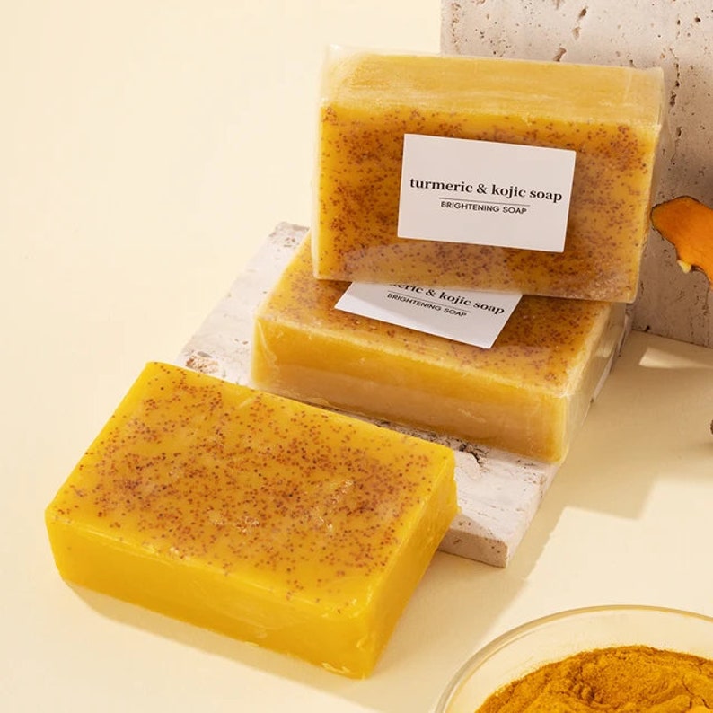 Puede incluir: Tres barras de jab&oacute;n amarillo de c&uacute;rcuma y kojic, envueltas en pl&aacute;stico transparente, con el texto "turmeric & kojic soap BRIGHTENING SOAP" en la etiqueta. El jab&oacute;n est&aacute; sobre una superficie blanca.
