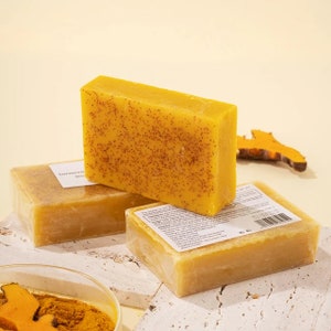 Puede incluir: Tres barras de jab&oacute;n amarillo de c&uacute;rcuma con una textura moteada. El jab&oacute;n est&aacute; apilado sobre una superficie blanca con un peque&ntilde;o taz&oacute;n de polvo de c&uacute;rcuma y un trozo de ra&iacute;z de c&uacute;rcuma.