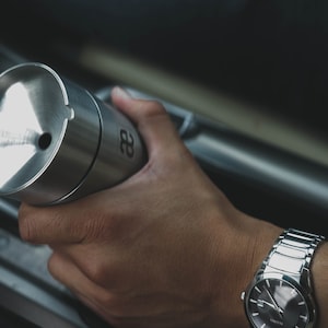 May include: A silver metal container with a black number "28" on the side. The container is being held by a person wearing a silver watch.