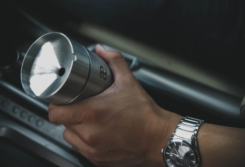 May include: A silver metal container with a black number "28" on the side. The container is being held by a person wearing a silver watch.