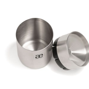 May include: A stainless steel coffee grinder with a black rubber base and a removable lid. The grinder has a logo that says "ae" on the side.