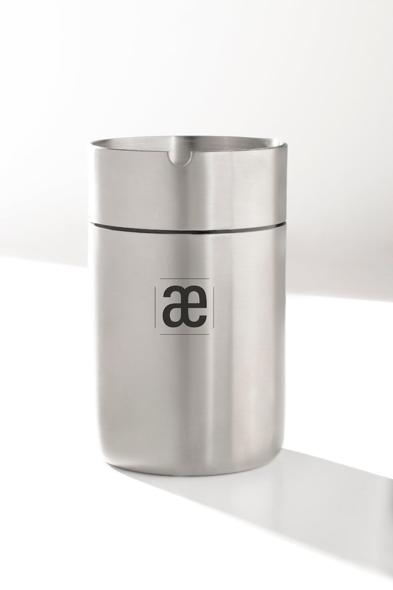 May include: A silver stainless steel container with a black logo that says "ae" inside a square.