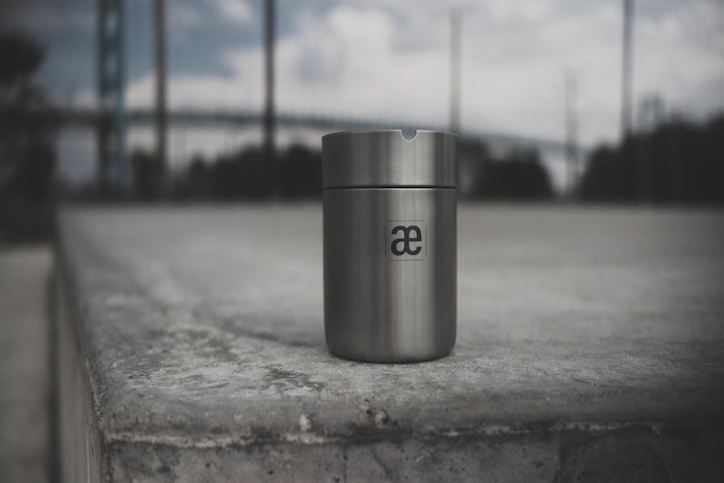 May include: A silver metal container with a lid and the letter "ae" printed on the side. The container is sitting on a gray concrete surface.