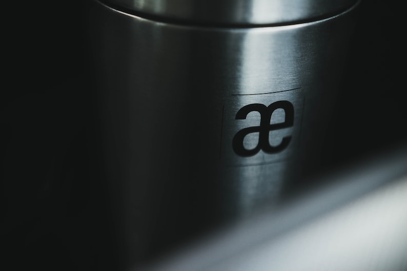 May include: Close-up of a stainless steel container with the letters "ae" printed on the side.