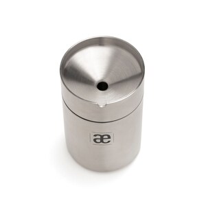 May include: A silver stainless steel container with a funnel-shaped top and a small hole in the center. The container has the letters "ae" printed on the side.