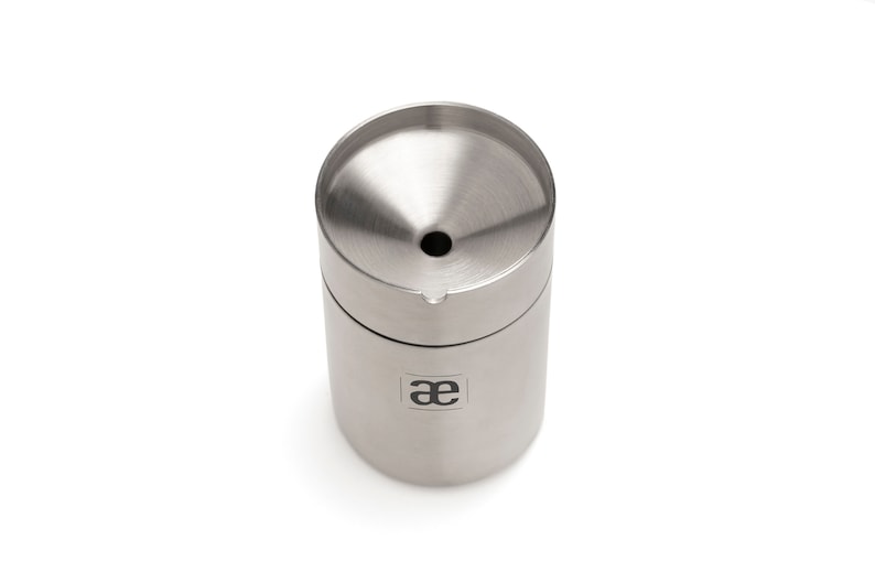 May include: A silver stainless steel container with a funnel-shaped top and a small hole in the center. The container has the letters "ae" printed on the side.