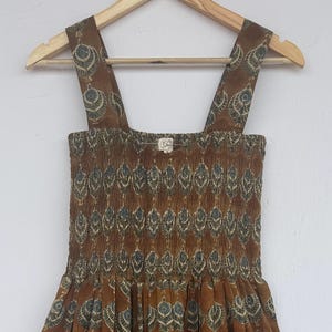 May include: A brown patterned sundress with wide straps. The dress features a smocked bodice and a tiered skirt. The fabric is a warm brown with a repeating pattern of stylized leaves in shades of blue and green.