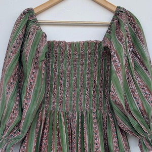 Może przedstawiać: Zielono-czerwona sukienka w paski z kwadratowym dekoltem i długimi rękawami. Sukienka ma dopasowany gorset i zwiewną spódnicę z falbanką. Tkanina ma pionowy wzór w paski z kwiatowymi akcentami.