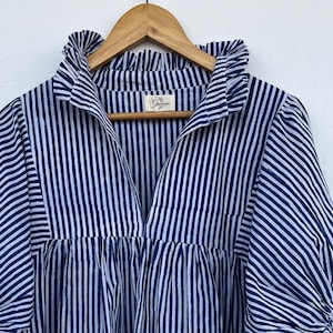 May include: A blue and white striped top with a frilled collar and sleeves. The top is hanging on a wooden hanger. The fabric has vertical stripes and a V-neckline. The sleeves are gathered at the cuffs.