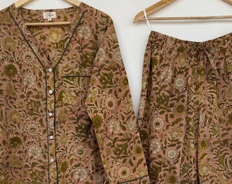 May include: A two-piece pajama set in a floral pattern. The set includes a long-sleeved button-down shirt and matching pants. The fabric has a pinkish-brown base color with a green and off-white floral design. The shirt has a V-neck and green trim.