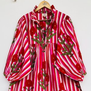 May include: A long, striped dress in red and white, with a floral pattern. The dress has long, puffy sleeves and a ruffled collar. The floral design is in red and green, contrasting with the striped background. The dress is displayed on a wooden hanger.