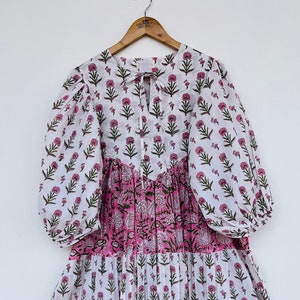 Könnte beinhalten: Ein weißes Kleid mit rosa Blumenmuster, einem Stufenrock und Puffärmeln. Das Kleid hat einen V-Ausschnitt und eine Schleife am Ausschnitt.