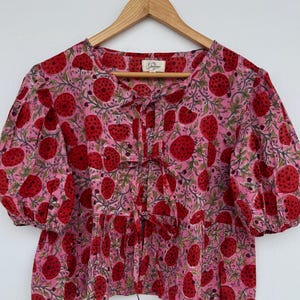 Op de afbeelding: Roze blouse met een rood aardbeienpatroon. De top heeft korte, pofmouwen, een strikhalslijn en een geplooid zoom. De stof is lichtroze met een herhalend patroon van rode aardbeien en groene bladeren.